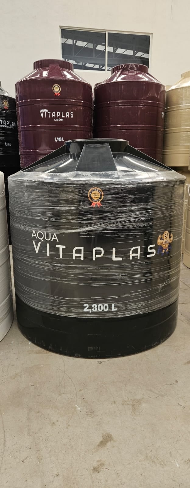 Large cylindrical Aqua Vitaplas water storage tank, 2,300L capacity, wrapped in protective plastic in an industrial storage facility with larger tanks visible behind it
